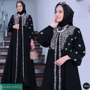 May include: Black abaya with floral embroidery and a long, flowing silhouette. The abaya has a high neckline and long sleeves. The model is wearing a black headscarf and sunglasses.