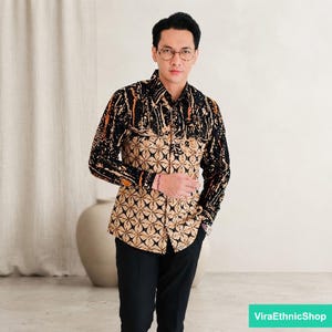 May include: A long-sleeved button-down shirt featuring a black and tan batik pattern. The upper chest and sleeves display a dark, abstract splatter design, while the lower torso showcases a repeating geometric star motif in tan and black.