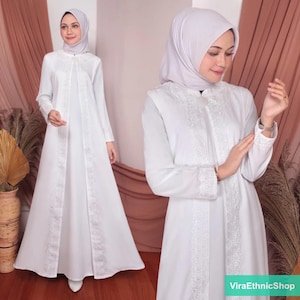 May include: A white abaya with long sleeves and a lace trim. The abaya has a button closure at the neck and a flowing skirt. The model is wearing a white headscarf.