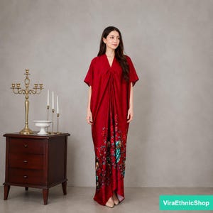 May include: A floor-length, deep red kaftan dress featuring a V-neckline and short sleeves. The garment is adorned with a vibrant floral and bird print concentrated along the lower front section, showcasing shades of blue, green, and white.