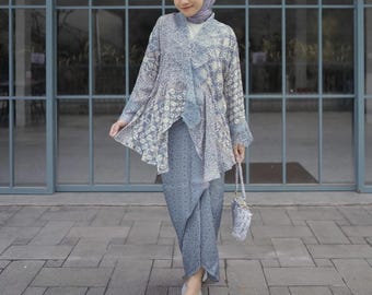 May include: A woman wearing a light blue patterned top with a matching skirt and a light blue headscarf. She is holding a small light blue purse with a beaded strap. The woman is standing in front of a glass door.