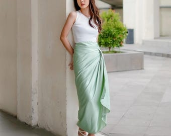 May include: A woman wearing a white tank top and a light green satin wrap skirt. She is standing next to a white wall and looking to the side.
