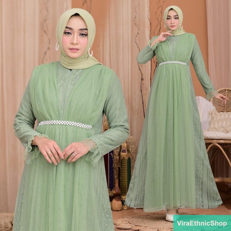May include: A long, flowing green maxi dress with delicate lace detailing and a pearl-embellished waist band. The dress has long sleeves and a modest neckline.