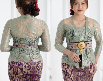 May include: A woman is wearing a sage green lace top with long sleeves and a matching skirt. She is wearing a gold and black belt with a large gold buckle. The skirt has a purple and gold floral pattern.