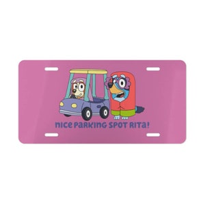 May include: A pink license plate with a cartoon dog driving a toy car and another cartoon dog standing next to the car. The text on the license plate reads "nice parking spot Rita!"