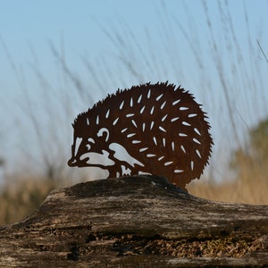 May include: A rusty metal silhouette of a hedgehog, standing on a weathered log. The hedgehog is facing left and has a detailed design with spikes and a snout.