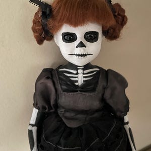 May include: A handmade doll with a skeleton face and body paint. The doll has red hair styled in two buns, and wears a black dress with a lace skirt. The doll's arms and legs are painted to resemble a skeleton.