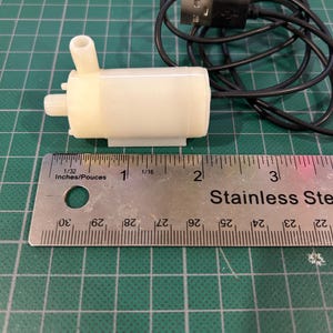 Può includere: Una piccola pompa per acqua sommergibile bianca con un cavo USB nero. La pompa è lunga circa 2,5 cm e viene misurata con un righello.