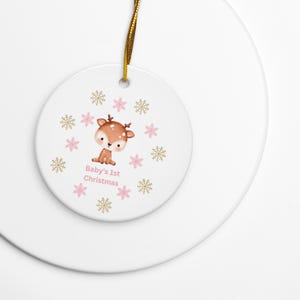 Può includere: Un ornamento rotondo in ceramica bianca con un'illustrazione di un cerbiatto a cartone animato marrone circondato da fiocchi di neve dorati e rosa. Il testo Baby's 1st Christmas è stampato in rosa sotto il cerbiatto. Un filo dorato è fissato in alto.