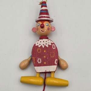 May include: A wooden pull-string toy clown with a red and white striped hat and a red nose. The clown has a red body with white polka dots and yellow feet. The arms are beige and the face is painted with blue accents.