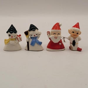 Puede incluir: Cuatro pequeñas figuras navideñas de cerámica. El conjunto incluye un muñeco de nieve con sombrero negro y bufanda azul, un muñeco de nieve con sombrero negro y una escoba, Papá Noel con traje rojo y un elfo sonriente con gorro rojo.