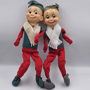Puede incluir: Dos muñecos de duende vintage con caras sonrientes, que visten pantalones rojos, tops verdes y grises y bufandas blancas. Los muñecos tienen zapatos y sombreros grises y verdes. Los muñecos posan como si estuvieran bailando.