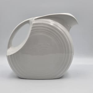 May include: A white ceramic pitcher with a rounded body and a curved handle. The pitcher has a spout and is decorated with concentric ridges. The design is simple and elegant, suitable for serving beverages.