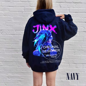 Può includere: Felpa con cappuccio blu navy con una grafica di un personaggio e la parola "JINX" in rosa e viola. La parte posteriore della felpa presenta anche il testo "ORIGINAL DESIGNER" e "designed by Rena".
