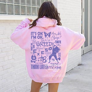 Könnte beinhalten: Ein hellrosa Kapuzenpullover mit verschiedenen Grafiken und Text in dunklerem Lila. Auf der Rückseite des Hoodies stehen Sätze wie "IT'S OK, I'M OK" und "Are We Flying?" sowie ein Schmetterling und andere Designs. Der Hoodie hat eine lockere Passform.
