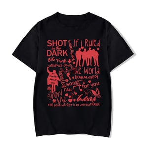 May include: Black t-shirt featuring red text and illustrations. The design incorporates song titles and phrases such as "Shot in the Dark" and "If I Ruled the World." Musical notes and silhouettes are also included.