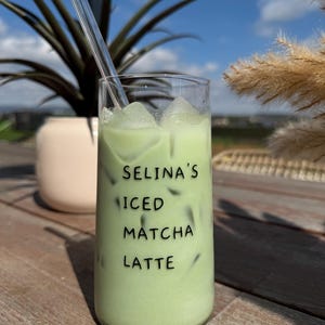 Könnte beinhalten: Ein Glas mit eisgekühltem Matcha-Latte mit Eiswürfeln. Auf dem Glas steht "Selina's Iced Matcha Latte".