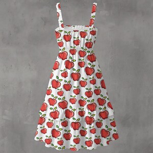 May include: White sundress with red apple pattern. The dress features a square neckline, thin straps, and a tiered skirt. The apples have green leaves and stems. The dress is made of a lightweight fabric.