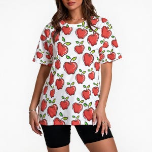 Puede incluir: Camiseta blanca con un estampado repetido de manzanas rojas con hojas verdes. La camiseta de manga corta se lleva con pantalones cortos negros. Las manzanas tienen contornos negros.