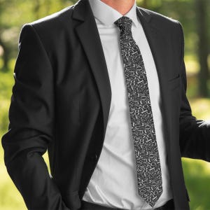 May include: A black suit with a white dress shirt and a patterned tie. The tie features a repeating pattern of numbers in shades of gray and white. The suit jacket is buttoned and the background is blurred.