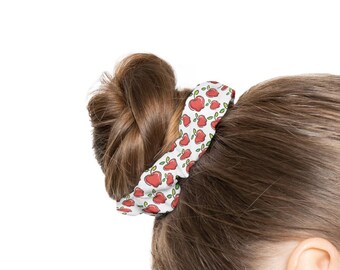 May include: A white scrunchie with a repeating pattern of red heart-shaped apples and green leaves. The scrunchie is wrapped around a bun of brown hair. The scrunchie is made of a soft fabric.