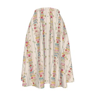May include: A cream-colored skirt featuring a repeating vertical pattern of stacked books in various pastel colors, interspersed with small red apples and thin blue stripes. The garment has an elasticized waistband and a flared, A-line silhouette.