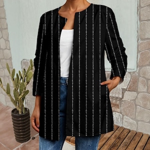 May include: Black open-front jacket with vertical white lines of numbers. The jacket has long sleeves and pockets. The person is wearing a white top and blue jeans. The jacket is a unique piece of clothing.