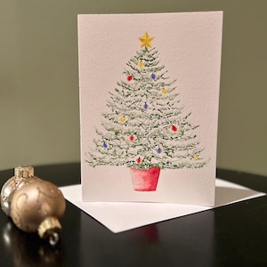 May include: Watercolor Christmas card featuring a decorated Christmas tree with a gold star on top. The tree is adorned with colorful lights and sits in a red pot. A gold and silver ornament is in the foreground.