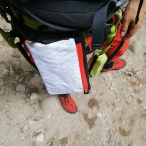 May include: A white and red zippered pouch with a diamond pattern is attached to a green and black hiking harness.
