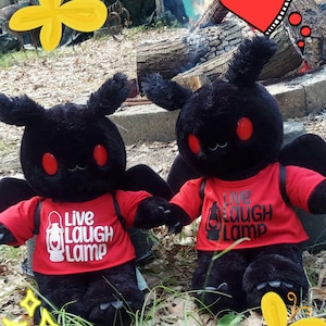May include: Two black plush moth toys with red eyes and small wings. Each wears a red t-shirt with the words "Live Laugh Lamp" printed in white. The toys are sitting in grass.