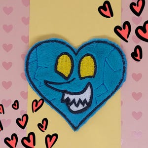 May include: A blue heart-shaped patch with a cartoonish face, featuring yellow eyes and a white-toothed grin. The patch has a textured appearance and is surrounded by a black border. Pink and red hearts are in the background.