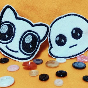 Peut inclure: Deux patchs brodés à repasser, l'un représentant un chat blanc aux grands yeux et un sourire, l'autre un fantôme blanc aux grands yeux et une moue.