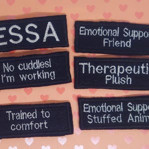 May include: Six black rectangular patches with white embroidered text. The patches read: ESSA, Emotional Support Friend, No cuddles! I'm working, Therapeutic Plush, Trained to comfort, and Emotional Support Stuffed Animal.