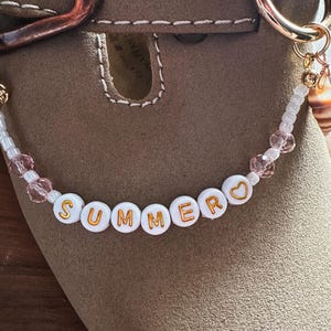 Peut inclure: Gros plan d'une chaussure beige avec un porte-clés doré et un charm en perles. Le charm indique "SUMMER" en lettres dorées sur des perles blanches, avec des perles roses et un coquillage.