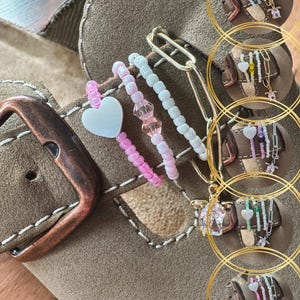Può includere: Primo piano di un sandalo in camoscio marrone con ciondoli decorativi di perline. I ciondoli includono perline rosa, bianche e trasparenti, un cuore bianco e una graffetta dorata. Il sandalo ha una fibbia color rame.