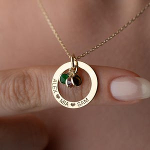 May include: A gold-colored necklace with a circular pendant. The pendant has the names "ALEX", "MIA", and "SAM" engraved on it. The necklace features three small, colorful charms and is worn around the neck.