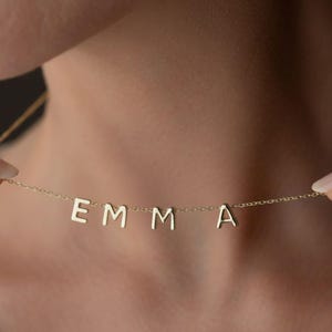 May include: A delicate gold necklace spelling out the name "EMMA" in capital letters. The pendant letters are evenly spaced and suspended from a fine chain. The necklace is shown against a neutral background.