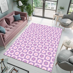 May include: A large rectangular area rug with a repeating floral pattern. The rug features a checkerboard design with alternating squares of light purple and pink. Each square contains a stylized flower in shades of pink and purple. The rug is placed on a light-colored wooden floor.