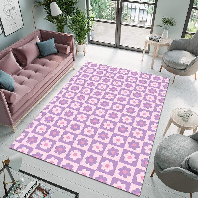 May include: A large rectangular area rug with a repeating floral pattern. The rug features a checkerboard design with alternating squares of light purple and pink. Each square contains a stylized flower in shades of pink and purple. The rug is placed on a light-colored wooden floor.