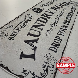 May include: A white sign with black text that reads "Self-Service 25 Loads of Laundry Room Open 24 Hours Drop Your Drawers Here". The sign has a distressed look and features decorative floral designs.