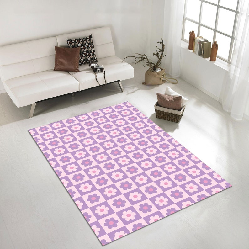 May include: A rectangular area rug with a repeating floral pattern. The rug features a checkerboard design with alternating squares of light purple and pink. Each square contains a white flower with a pink center. The rug is placed on a white floor.