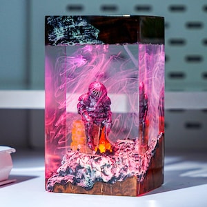 May include: A rectangular resin art piece with a miniature figure in a spacesuit, lit by pink and orange internal lighting. The base has a wood-like appearance, and the top has a dark, textured design.