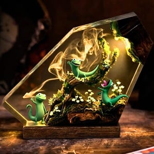 May include: A decorative lamp featuring three green gecko figurines on a branch inside a clear, geometric resin block. The lamp has a wooden base and emits a warm, yellow glow. A remote control with color options is visible.