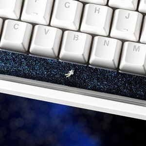 Può includere: Una barra spaziatrice per tastiera nera con un design a glitter blu e una piccola figura di astronauta.