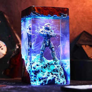 Op de afbeelding: Een rechthoekige hars sculptuur met een futuristisch soldatenfiguur. De figuur staat tegen een achtergrond van blauwe en witte wervelende patronen, met een houten boven- en onderkant. De sculptuur wordt verlicht met blauw licht.