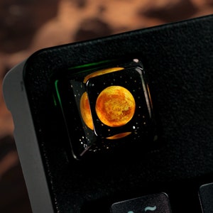 May include: A black keyboard keycap with a clear resin top featuring a yellow and orange planet with a black background.