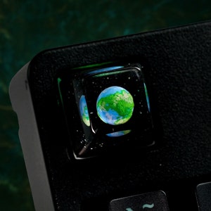 Könnte beinhalten: Ein grüner und blauer Erd-Tastenkappen mit schwarzem Hintergrund. Die Tastenkappe ist aus Harz und hat eine glänzende Oberfläche.