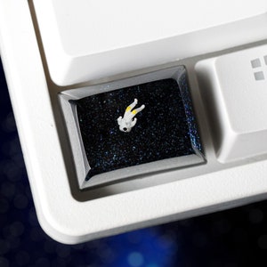 Peut inclure: Une figurine d'astronaute blanche avec des accents jaunes est placée sur un capuchon de touche noir avec une finition scintillante. Le capuchon de touche est sur un clavier blanc.