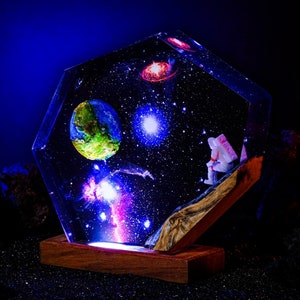 May include: A hexagonal resin sculpture depicting a galaxy with a blue nebula, a red nebula, a white star, and a green planet. A small astronaut figurine sits on a rocky surface in the foreground.