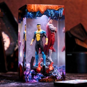 Puede incluir: Escultura rectangular de resina con dos figuras de superhéroes. Una figura viste un traje negro, azul y amarillo, y la otra una capa roja. La escultura tiene una base y una parte superior coloridas.